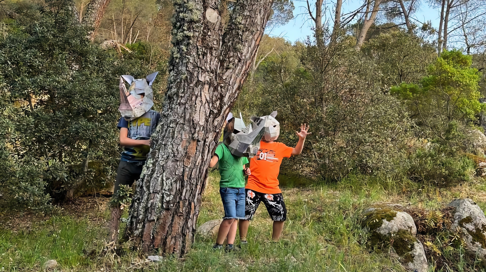 bambini con delle maschere in cartapesta giocano tra la vegetazione, vicino a un pino e delle rocce di graniti coperte di muschio.